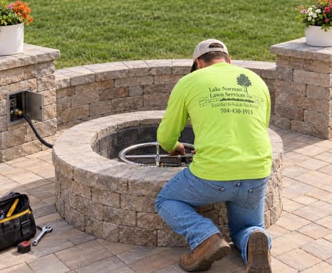 Crew member fitting a propane burner ring inside a block fire pit on a paver patio